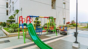 An outdoor playground with a green slide, swings, and a tire swing, surrounded by a landscaped garden and a building in the background. - Warm And Cozy Studio Apartment Margonda Residence 5 Near Campus (Depok)