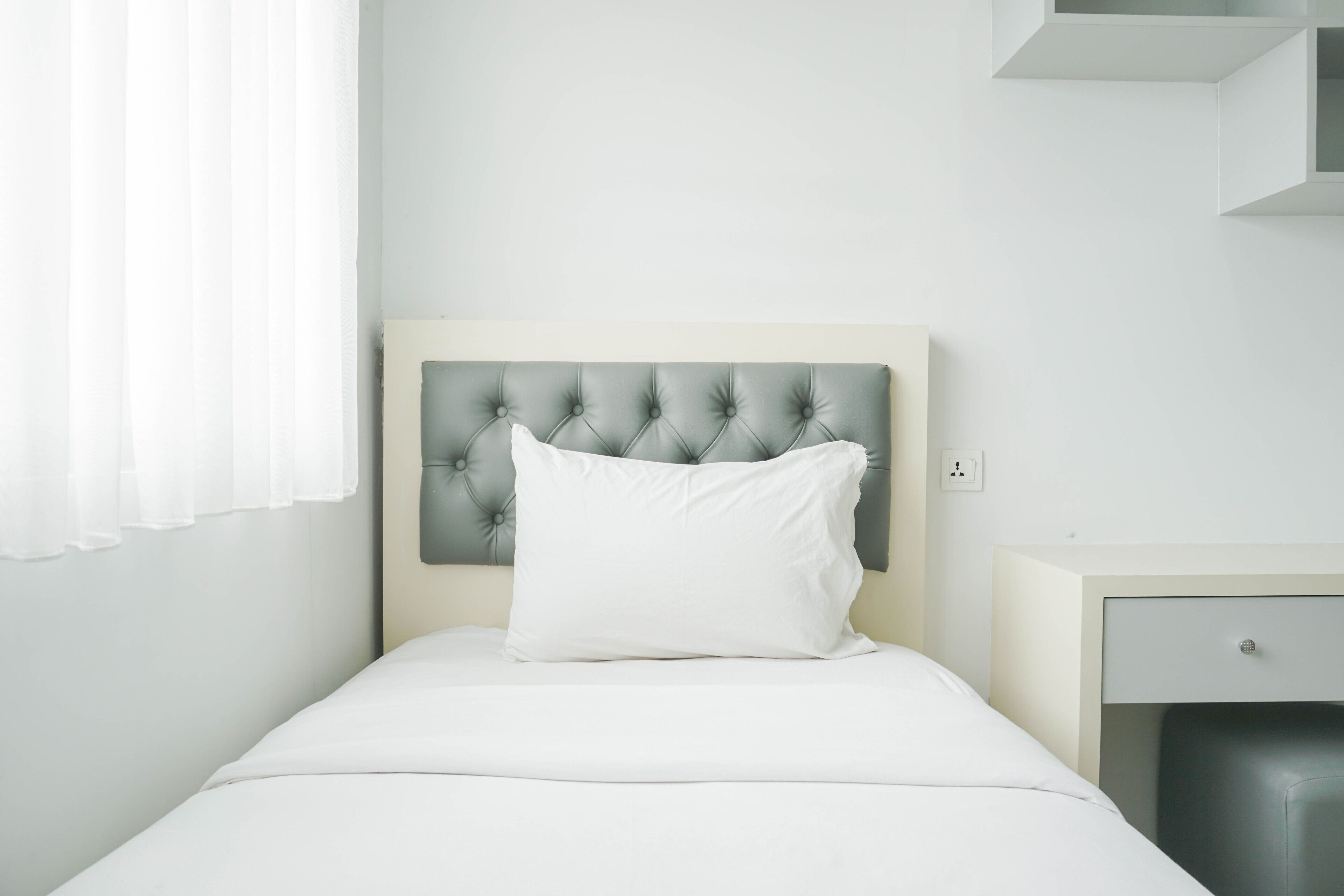 A neatly made bed with a tufted headboard, a white pillow, and a small bedside table with a drawer.