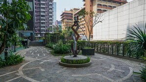 A courtyard with a central sculpture, surrounded by a patterned stone floor, greenery, and a building in the background.