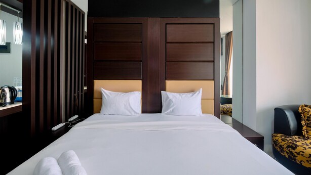 A hotel room with a large bed, two white pillows, a dark wooden headboard, a nightstand with a lamp, and a chair with a patterned seat. - Comfort And Homey Studio Apartment At Mangga Dua Residence (Jakarta)
