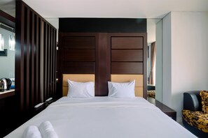 A hotel room with a large bed, two white pillows, a dark wooden headboard, a nightstand with a lamp, and a chair with a patterned seat. - Comfort And Homey Studio Apartment At Mangga Dua Residence (Jakarta)