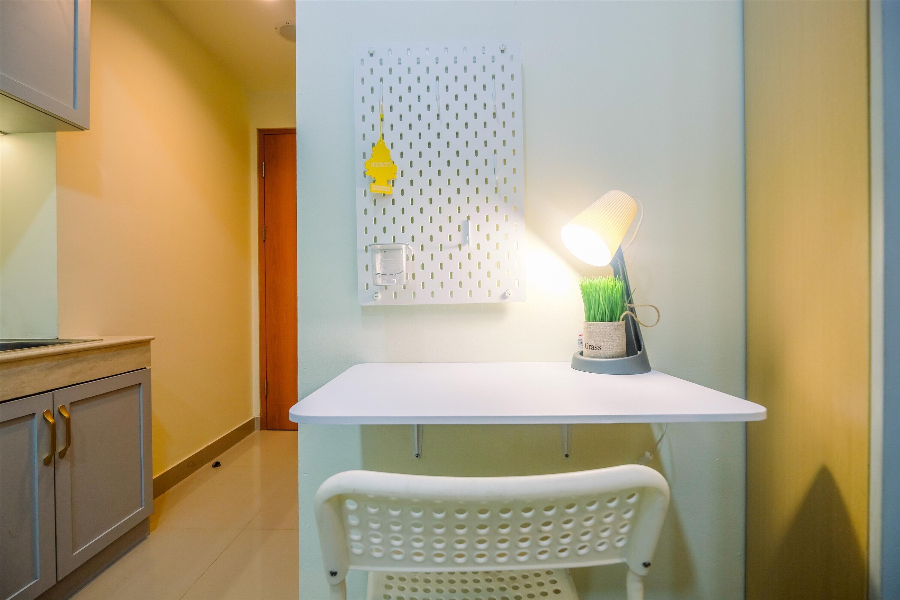A small kitchenette with a white countertop, a white chair, a hanging light fixture, and a wall-mounted shelf with a glass and a potted plant.