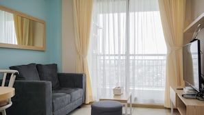 A modern living room with a grey sofa, a small round ottoman, a white table, and a TV stand with a flat-screen TV. - Comfortable And Nice 1Br At M-Town Signature Apartment (Gading Serpong)