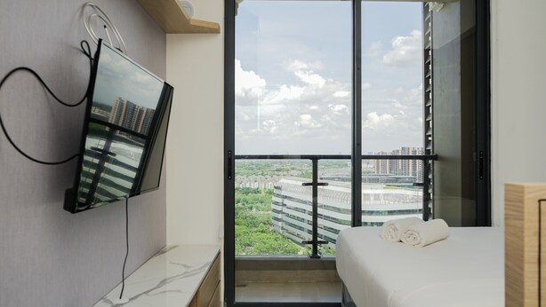 A modern hotel room with a bed, a TV mounted on the wall, and a balcony with a city view. - Comfortable And Simply Studio At Sky House Bsd Apartment (Pagedangan)