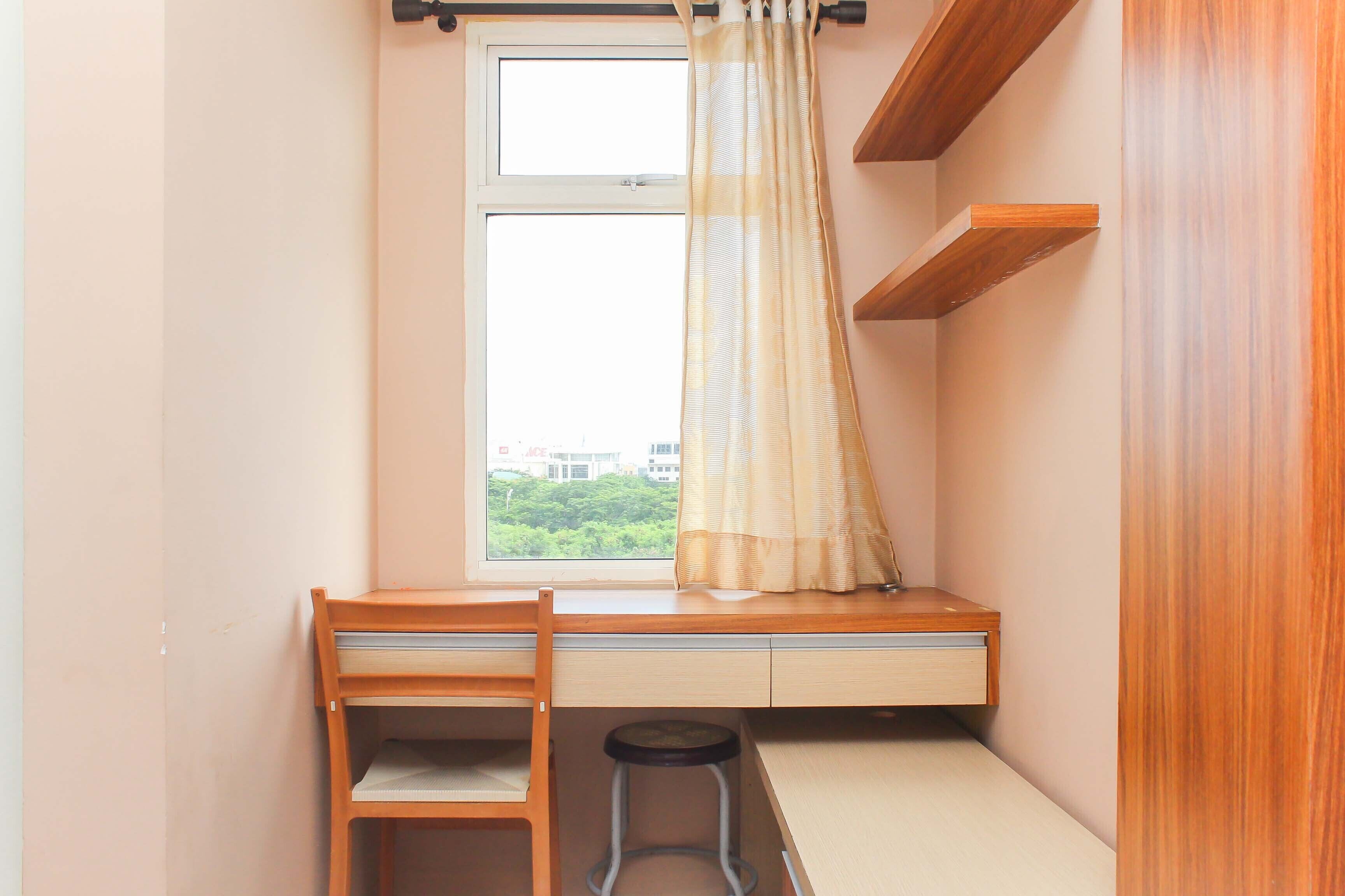 A small room with a desk, chair, and stool. There is a window with a curtain, and a view of greenery outside.