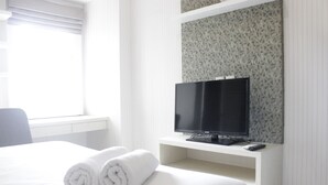 A hotel room with a bed, a TV on a stand, and a window with blinds. - Comfy & Scenic Studio Apartment At Parahyangan Residence Near Unpar (Bandung)