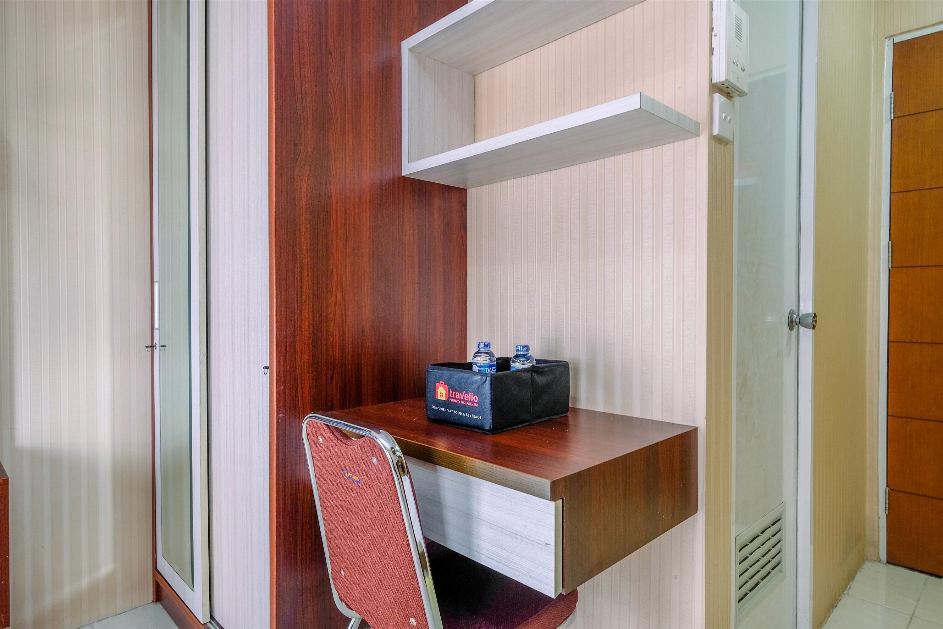 Quarto de hotel compacto com uma escrivaninha de madeira, uma cadeira, um minibar e um guarda-roupa.