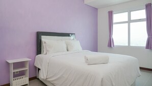 A neatly made bed with white linens and a rolled towel, a white nightstand with a shelf, and a window with purple curtains. - Comfort And Elegant 1Br Green Bay Condominium Apartment (Jakarta)