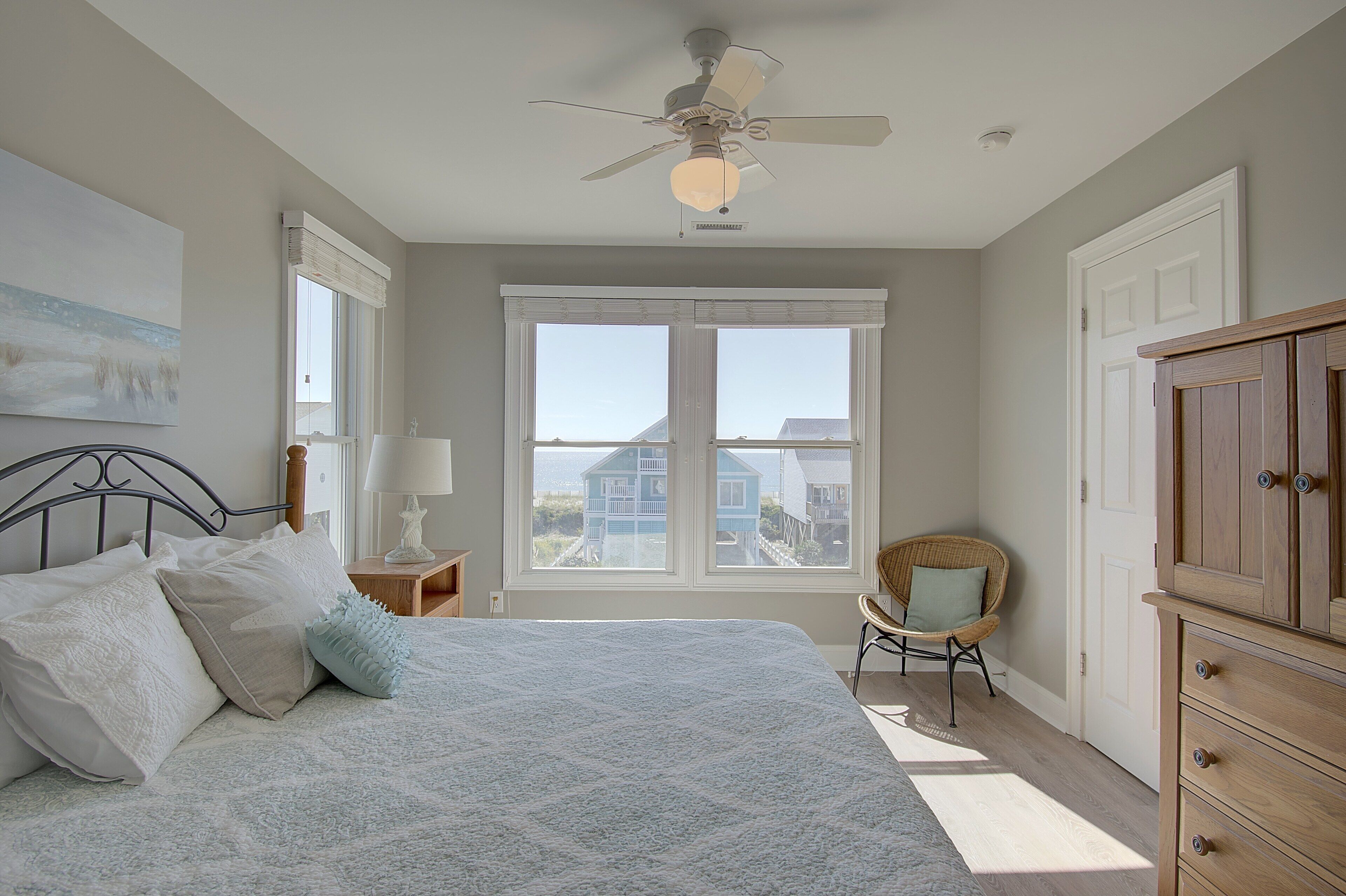A bedroom with a bed, a ceiling fan, a window with a view, a chair, and a wooden cabinet.