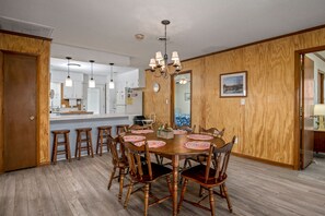 Zona pranzo con tavolo e sedie in legno, un lampadario e una cucina sullo sfondo.