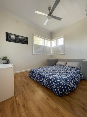 A bedroom with a bed, a ceiling fan, a window with blinds, and a painting on the wall. - Summer Breeze | 2BR. Parking. Close to CBD + Beach (The Junction)
