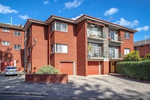 A two-story brick apartment building with a balcony on the second floor, a garage door, and a car parked in front. - Summer Breeze | 2BR. Parking. Close to CBD + Beach (The Junction)