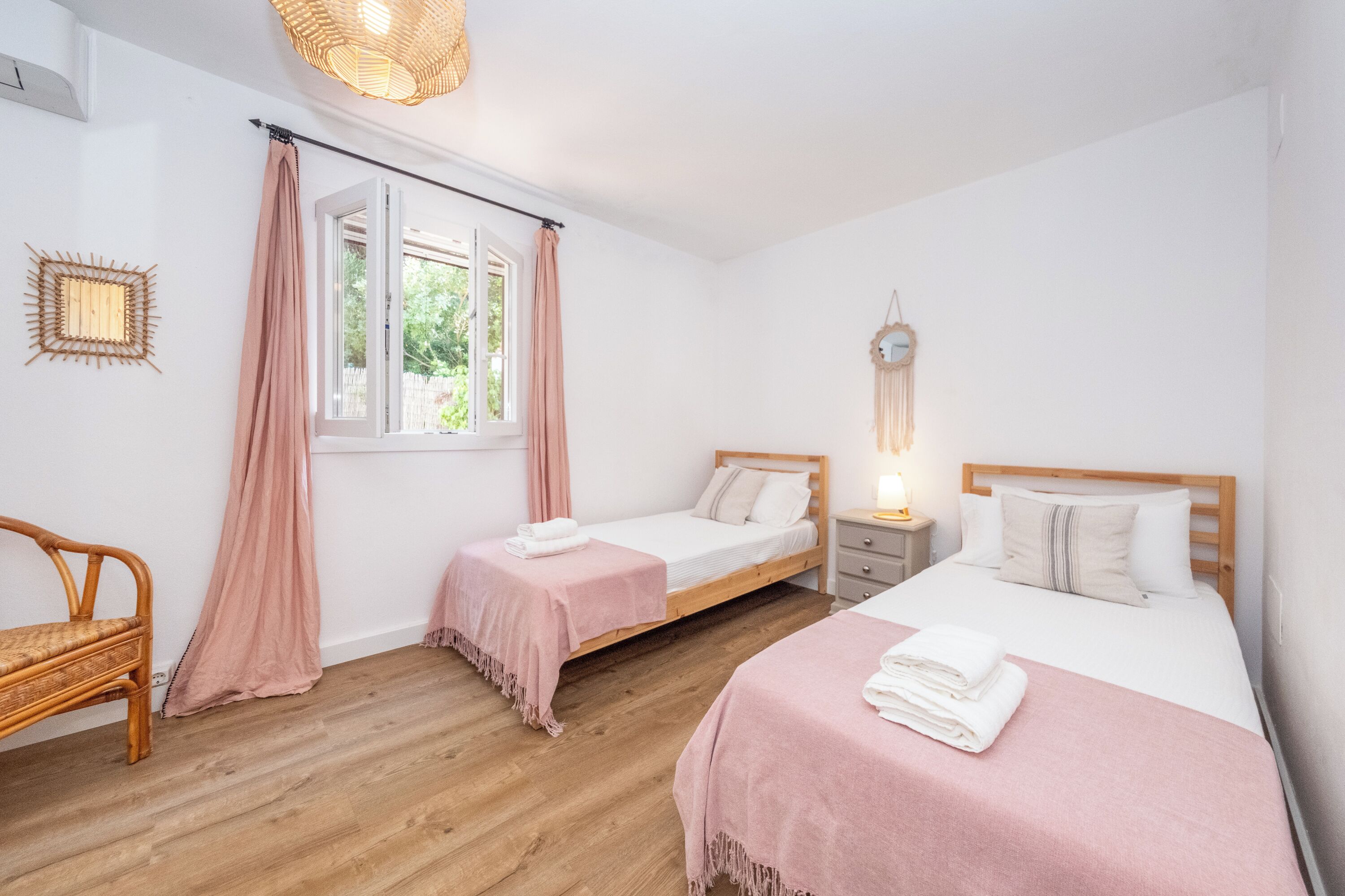 A bedroom with two beds, a window, a bedside table, a lamp, and a rattan chair.