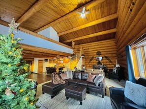 A spacious log cabin interior with a large wooden ceiling, a Christmas tree, a leather sofa, and a staircase leading to an upper level.