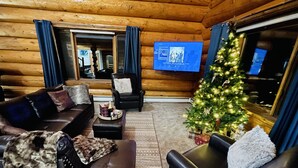 Interior acollidor d'una cabana de fusta amb un sofà de cuir, una taula de cafè amb una ampolla i gots, un arbre de Nadal i un televisió de pantalla plana penjada a la paret.