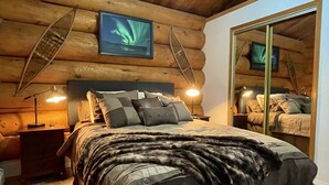 Un dormitori de cabana de fusta amb un llit, tauletes de nit, un mirall i un quadre amb l'aurora boreal.