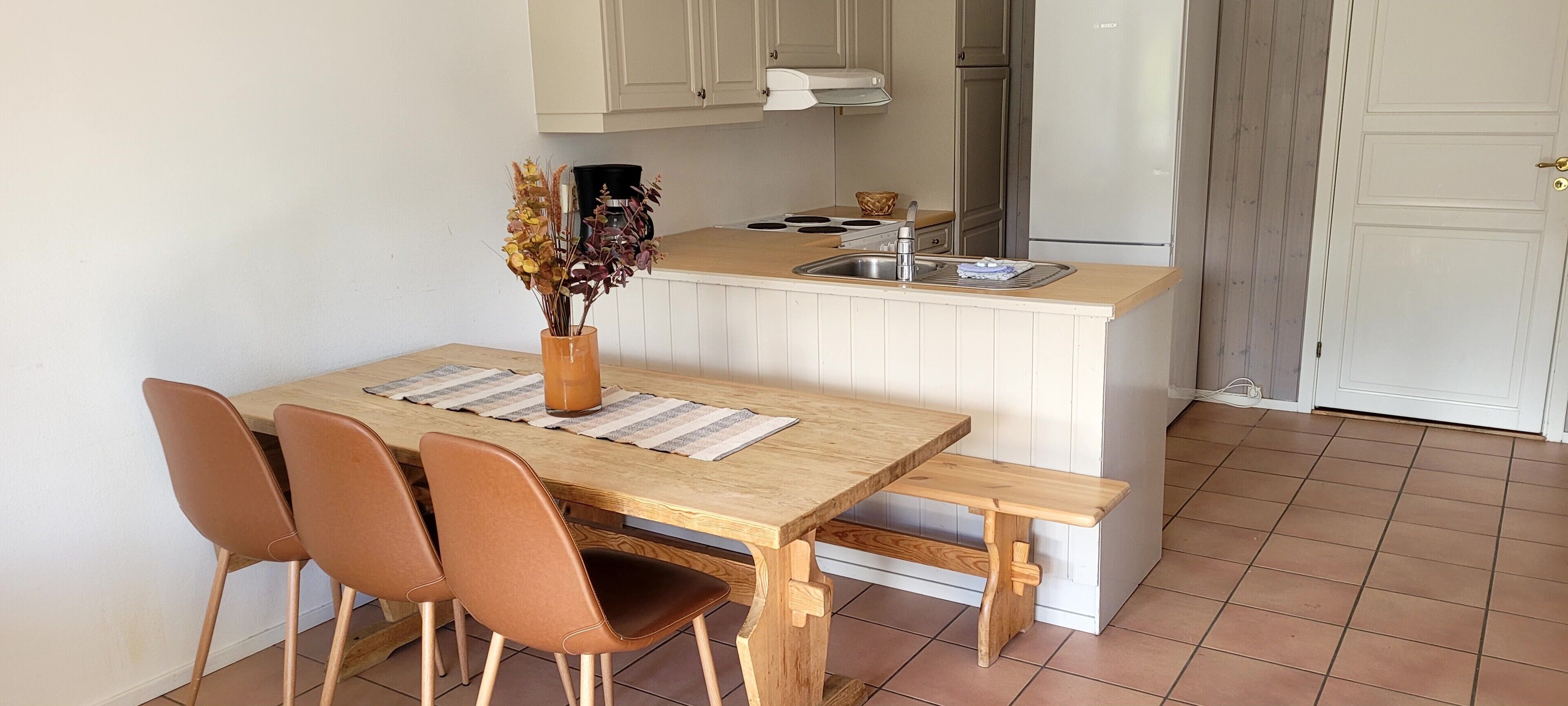 A compact kitchen with a wooden table, four chairs, a bench, a sink, and a refrigerator.