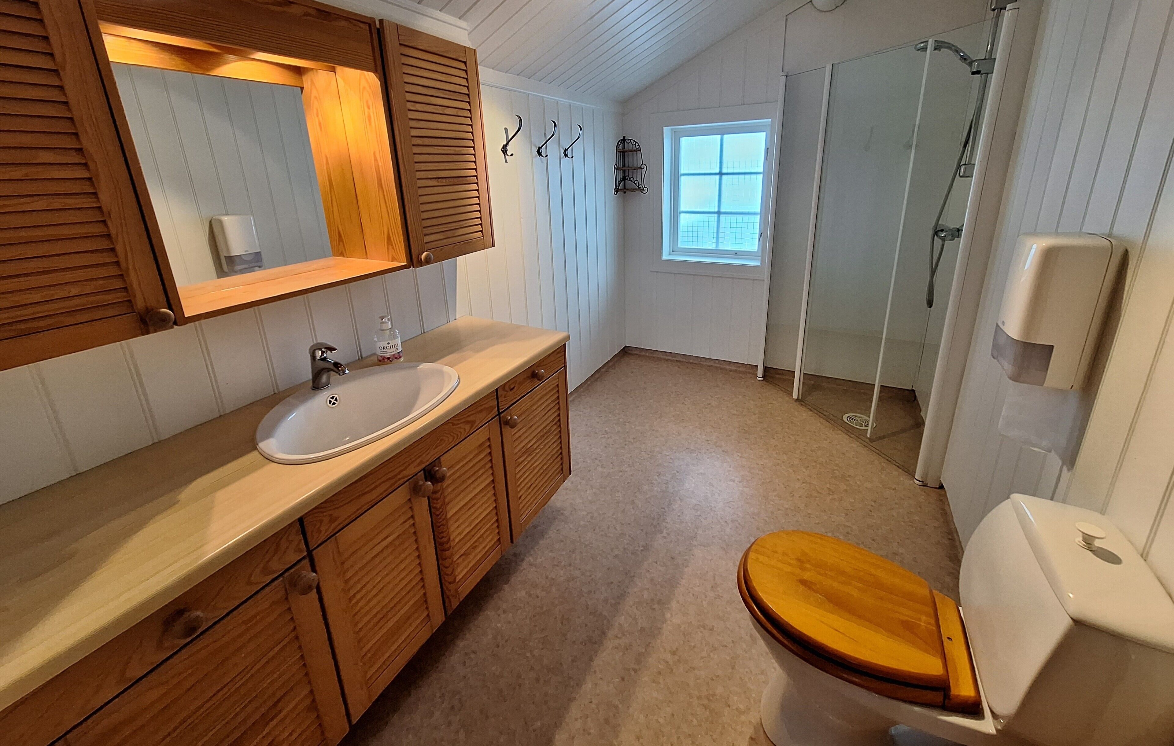 A bathroom with a wooden vanity, a white sink, a wooden toilet, a shower with a glass door, and a window with a view.