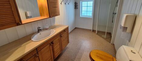 A bathroom with a wooden vanity, a white sink, a wooden toilet, a shower with a glass door, and a window with a view.
