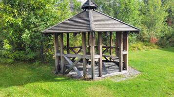 Un kiosque en bois doté d’un toit triangulaire et de côtés ouverts, entouré de pelouse et d’arbres.