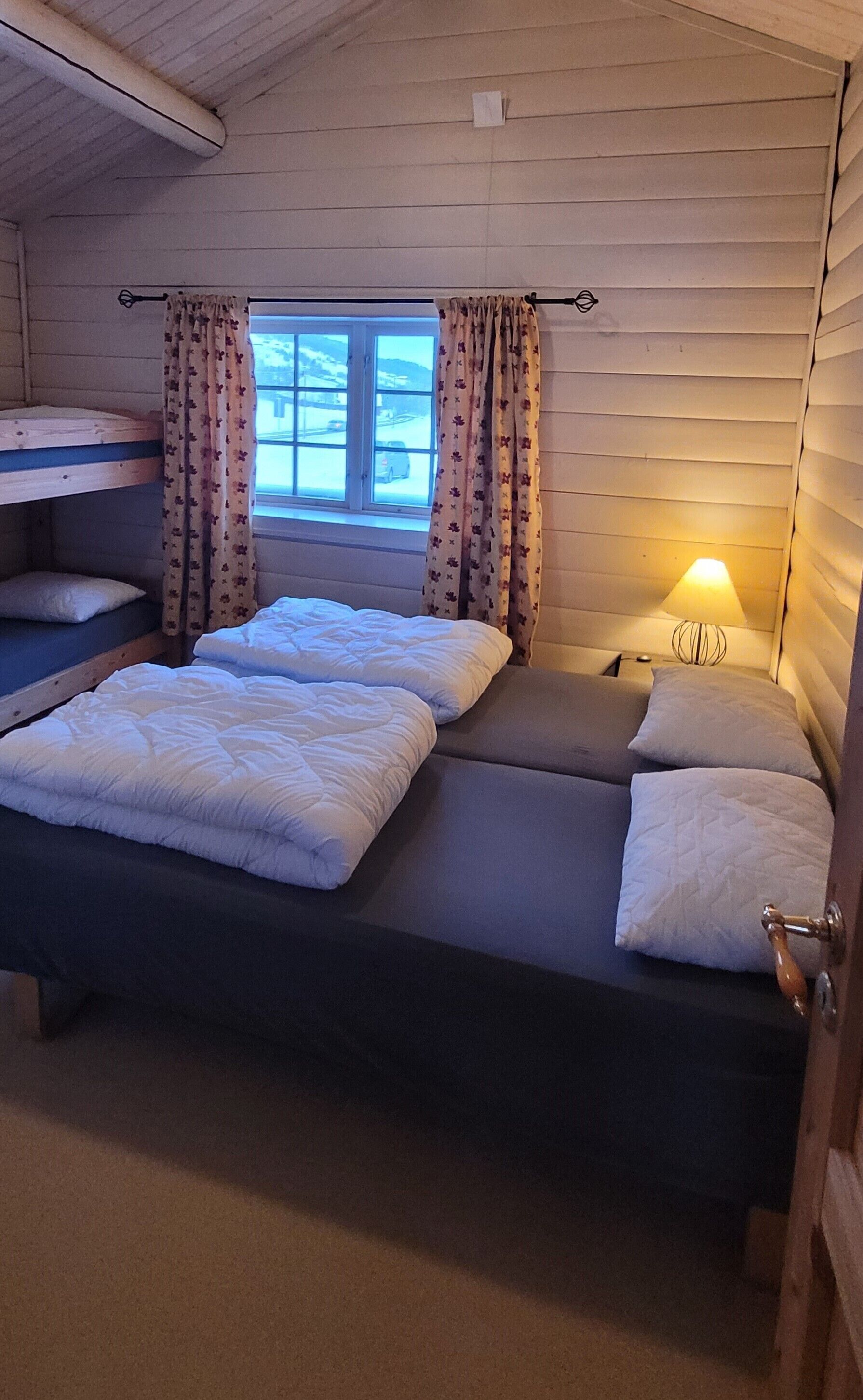 Ein kleines, gemütliches Zimmer mit einem Etagenbett und einem Einzelbett. Es gibt ein Fenster mit Vorhängen, eine Nachttischlampe und eine Holztür.