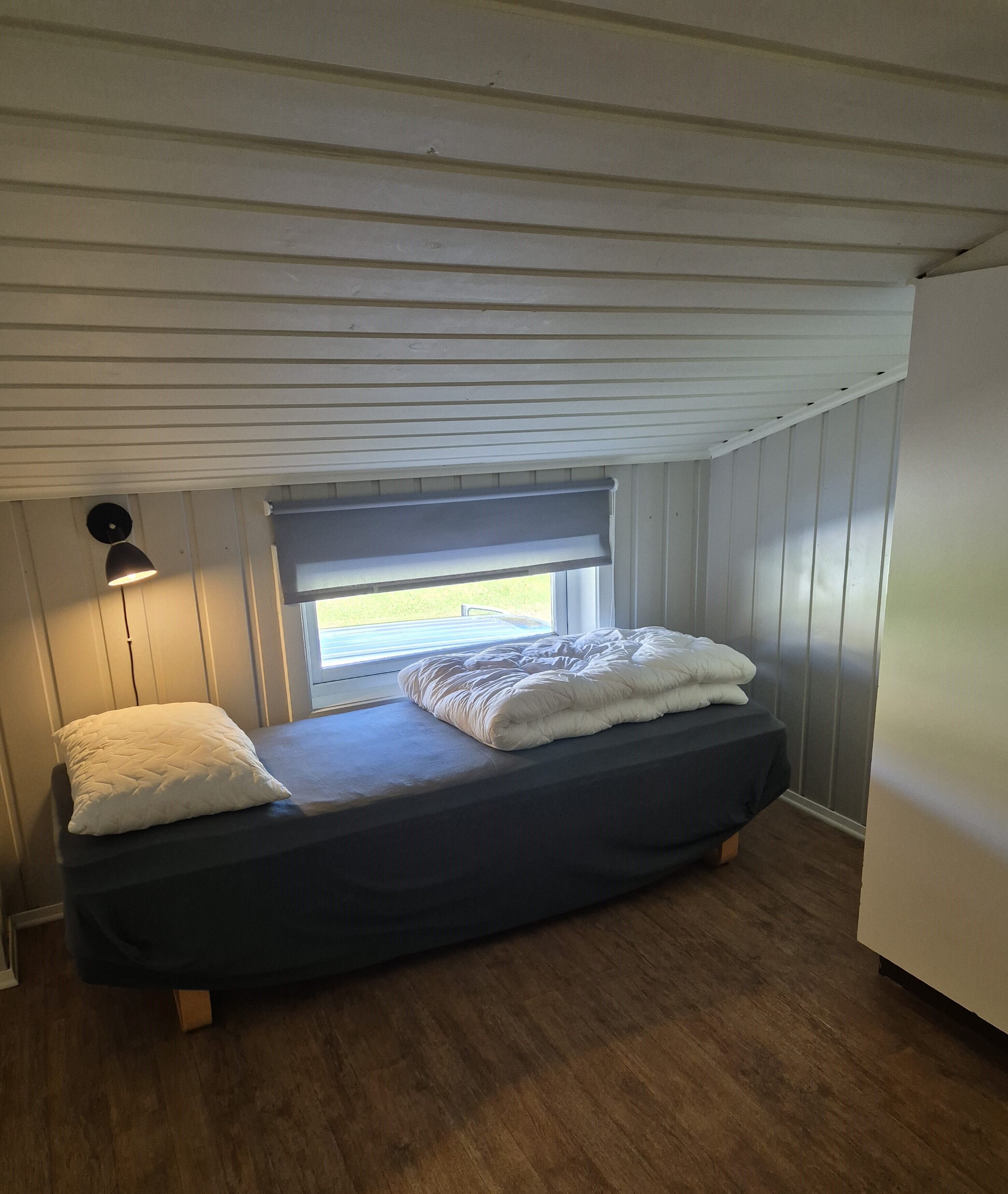 A small, single-bed room with a window, a bedside lamp, and a wooden floor.