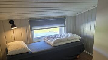 A small, single-bed room with a window, a bedside lamp, and a wooden floor.