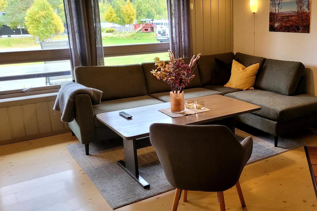En hyggelig stue med en sofa, et sofabord og en lænestol. Der er et vindue med udsigt, en pejs med murstensdesign, og et maleri på væggen.