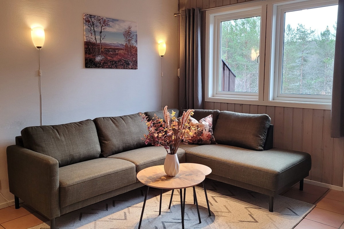 En stue med sofa, lille rundt bord, vase med visne blomster, et billede på væggen og et vindue med gardiner.