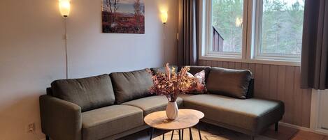 A living room with a sofa, a small round table, a vase with dried flowers, a painting on the wall, and a window with curtains.