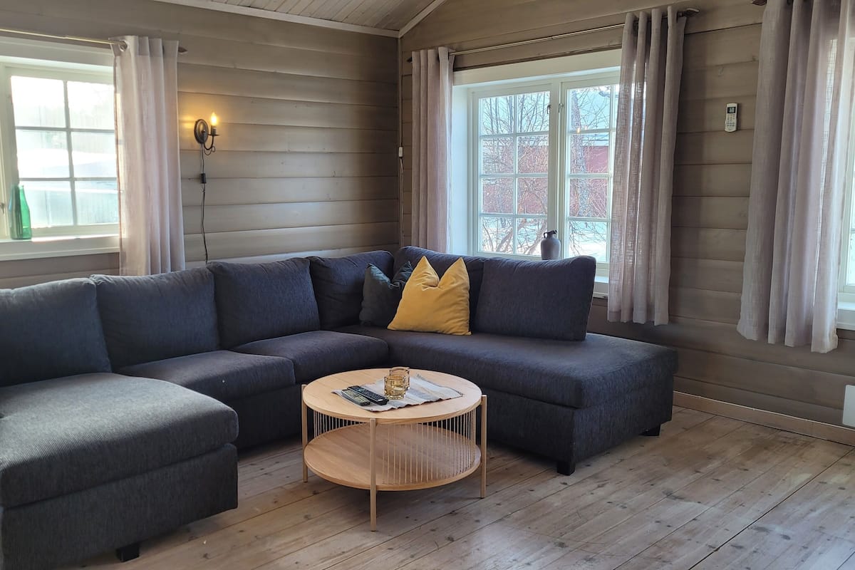 En moderne stue med en hjørnesofa, et rundt sofabord og store vinduer med gardiner.