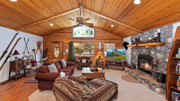 A cozy lodge interior with a wooden ceiling, a stone fireplace, a television, and a fireplace mantel with decorative items.