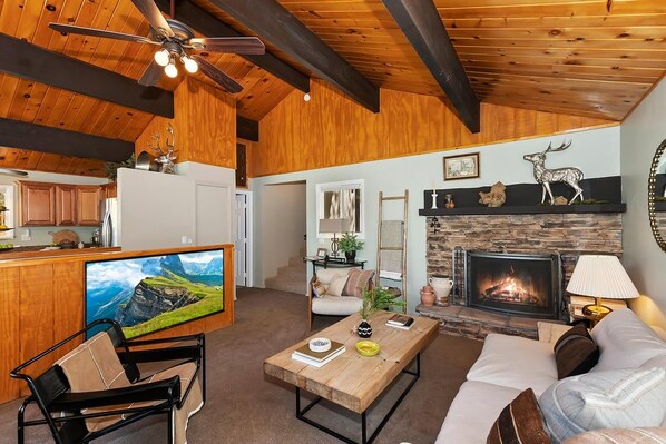 A cozy living area with a fireplace, wooden ceiling beams, a television, and a sofa. - Wonderful Moonridge Location! Mountain Views! Hot Tub! Pool Table! Spacious Deck! (Big Bear Lake)