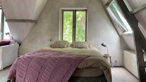 A bedroom with a sloped ceiling, a large window, a bed with a blanket, a bedside table, and a chair. - Mediterranean apartment of Guesthouse In de Aap - Finca - Hartje Bergen (Bergen (NH))
