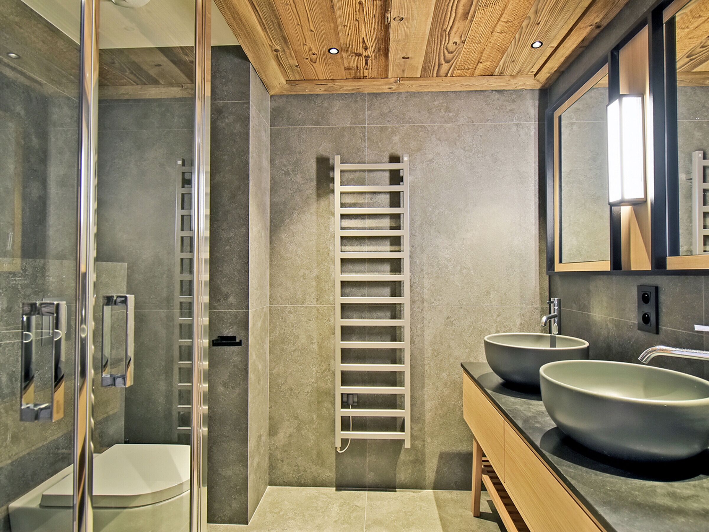 A modern bathroom with a glass shower enclosure, a white toilet, a wooden ladder shelf, and two sinks with a large mirror above.