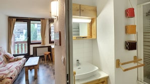Un espace de vie compact comprenant un canapé, une petite table, un meuble en bois, un évier blanc et un miroir placé au-dessus.