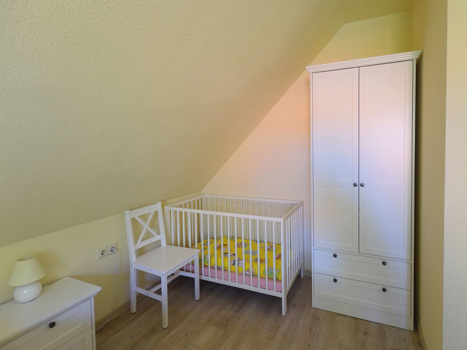 A room with a white crib, a white chair, a white nightstand, and a white wardrobe.