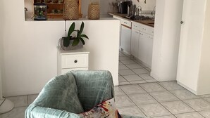 Un salon avec un fauteuil bleu, une petite table blanche ornée d’une plante, et un coin cuisine avec des meubles blancs et une cuisinière.