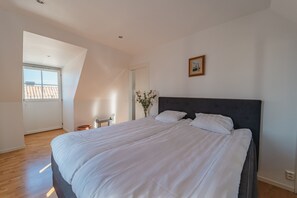 Een slaapkamer met een groot bed, een klein tafeltje, een stoel, een vaas met bloemen en een raam met uitzicht op gebouwen.