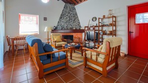 A living room with a stone fireplace, wooden furniture, a red door, and a tiled floor. - Cottage House with A/C – Big Garden - FREE Wood (Praia da Vitória)