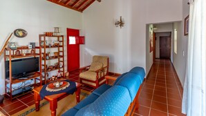 A living room with a television, wooden furniture, a red door, and a hallway. - Cottage House with A/C – Big Garden - FREE Wood (Praia da Vitória)