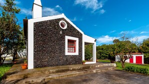 A small building with a stone facade, a red window, and a white door. - Cottage House with A/C – Big Garden - FREE Wood (Praia da Vitória)