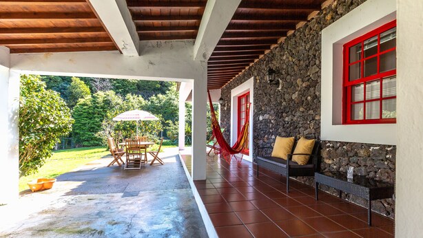 A covered patio with a stone wall, red-framed windows, and a hammock. - Cottage House with A/C – Big Garden - FREE Wood (Praia da Vitória)