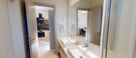 Une salle de bain moderne avec deux lavabos, un grand miroir et une vue sur la cuisine.