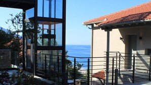 Una estructura moderna de vidrio con vista al mar, un patio de piedra y un edificio con techo de tejas.