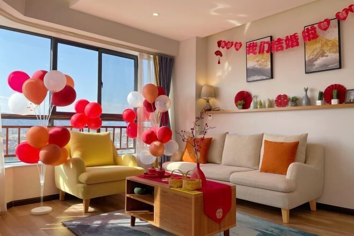 Una sala de estar moderna con un sillón amarillo, un sofá beige y una mesa de centro de madera. Hay globos en un soporte y un jarrón rojo con ramas. La habitación tiene ventanas grandes y vistas al exterior.