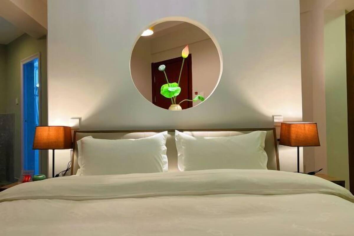 Ein Hotelzimmer mit einem Bett, zwei Nachttischlampen, einem runden Spiegel an der Wand und einer Tür links.