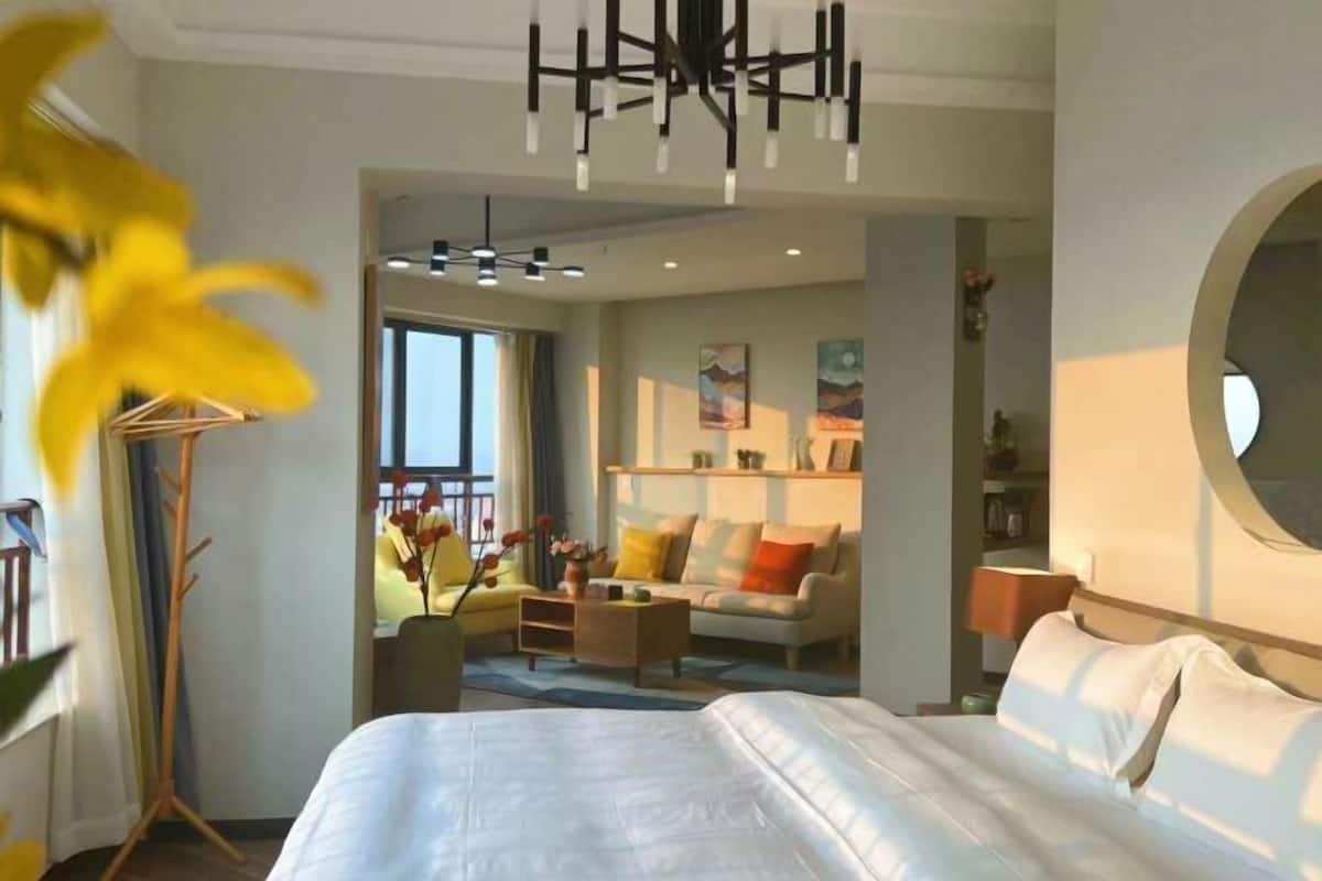Ein modernes Hotelzimmer mit einem großen Bett, einer Sitzecke mit Sofa und Sesseln, einem Couchtisch und einem Esstisch. Ein großes Fenster sorgt für natürliches Licht und bietet einen Ausblick ins Freie.