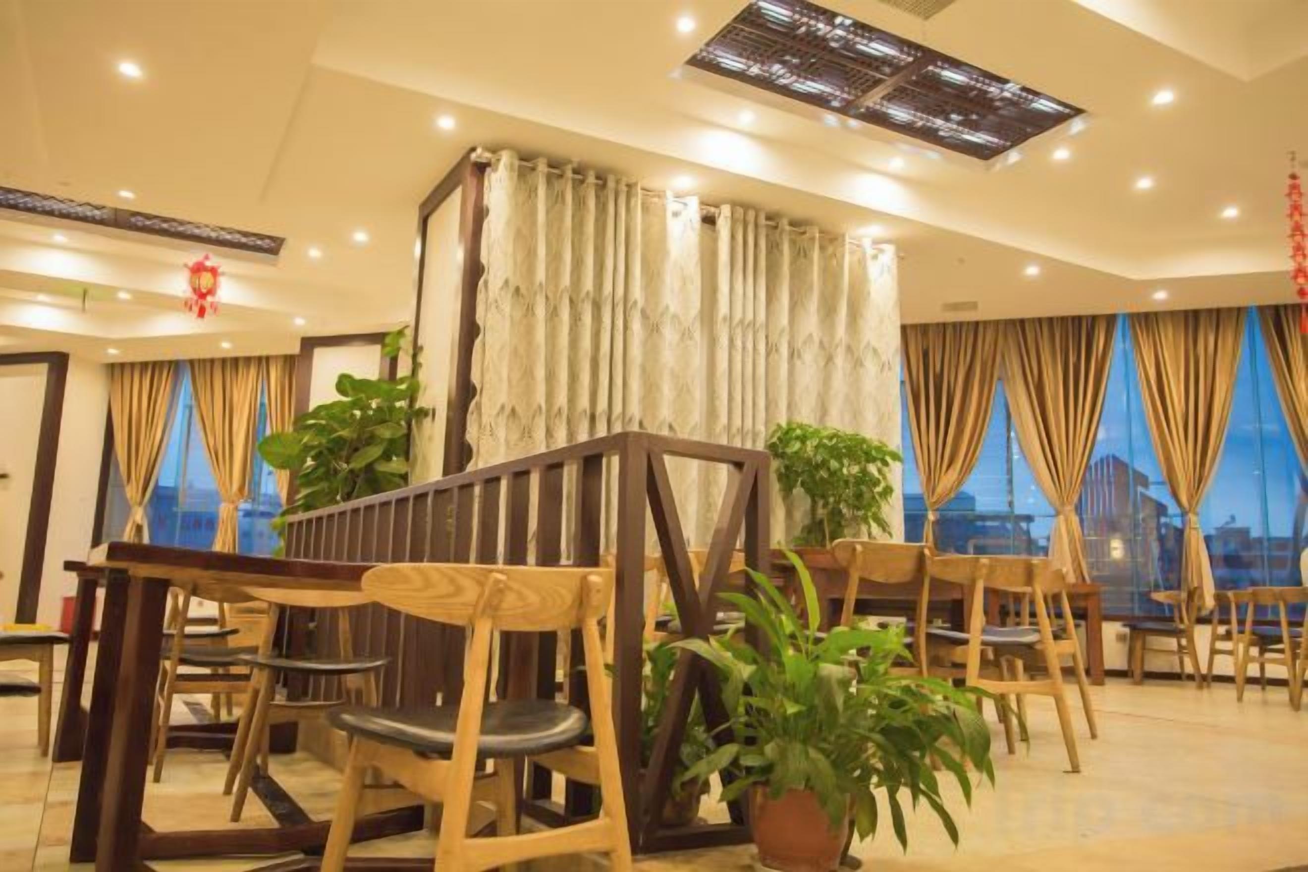 A hotel dining area with wooden furniture, potted plants, and large windows.
