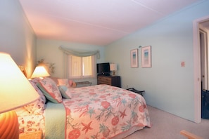 A bedroom with a bed, bedside lamps, a dresser, a TV, and a window with curtains. - Traditional, stylish 2 bedroom oceanfront condo with free WiFi, and outdoor pool, and a stunning ocean view located in quiet midtown near mini golf and bowling and just steps from the beach! (Ocean City)